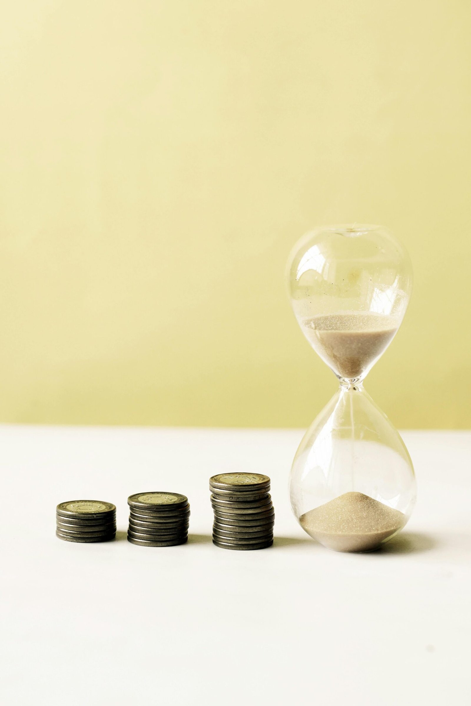 A vertical close-up of coins and an hourglass against a yellow background symbolizing time and money.