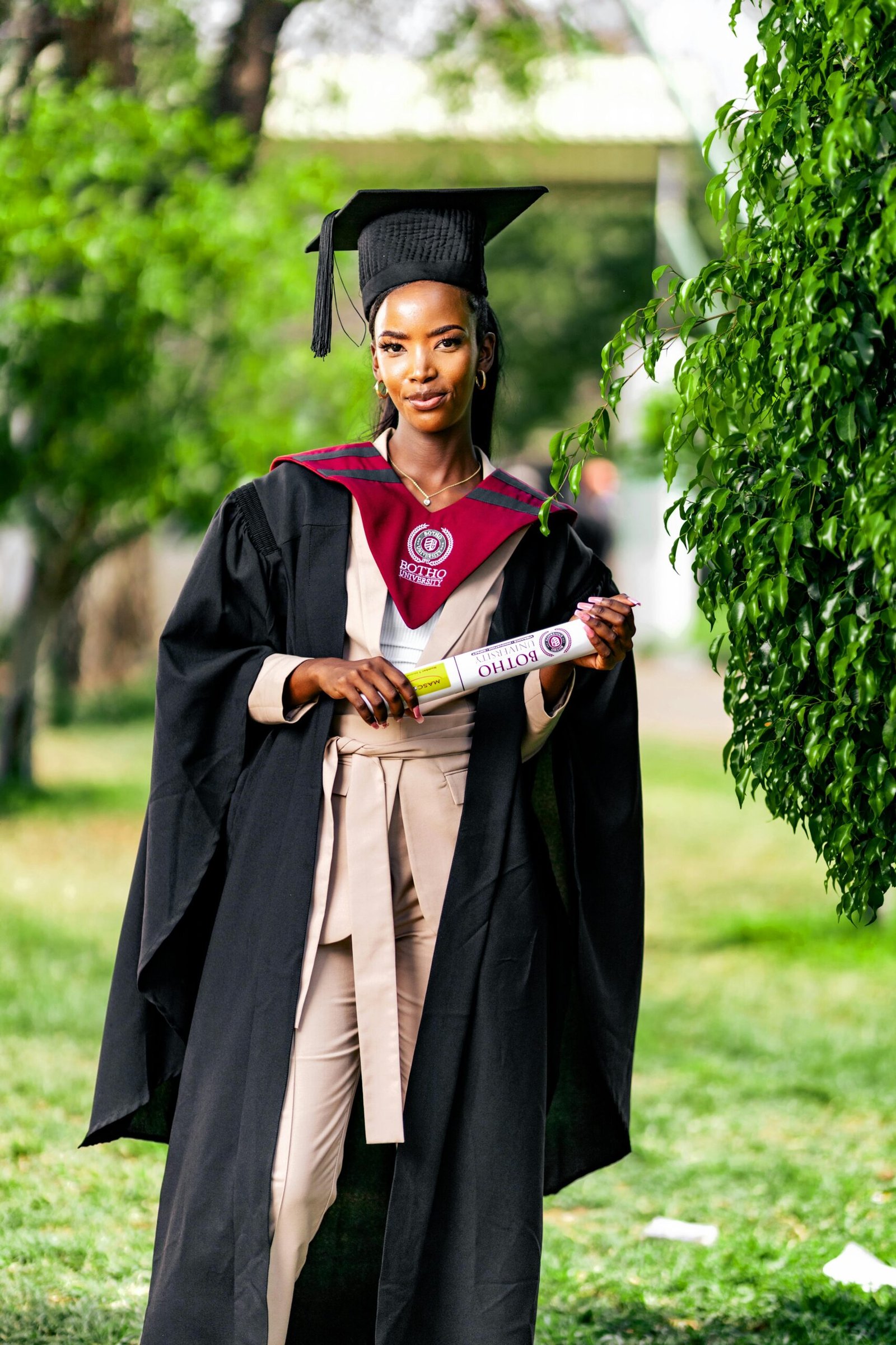 Proud female graduate in cap and gown holding diploma outdoors.