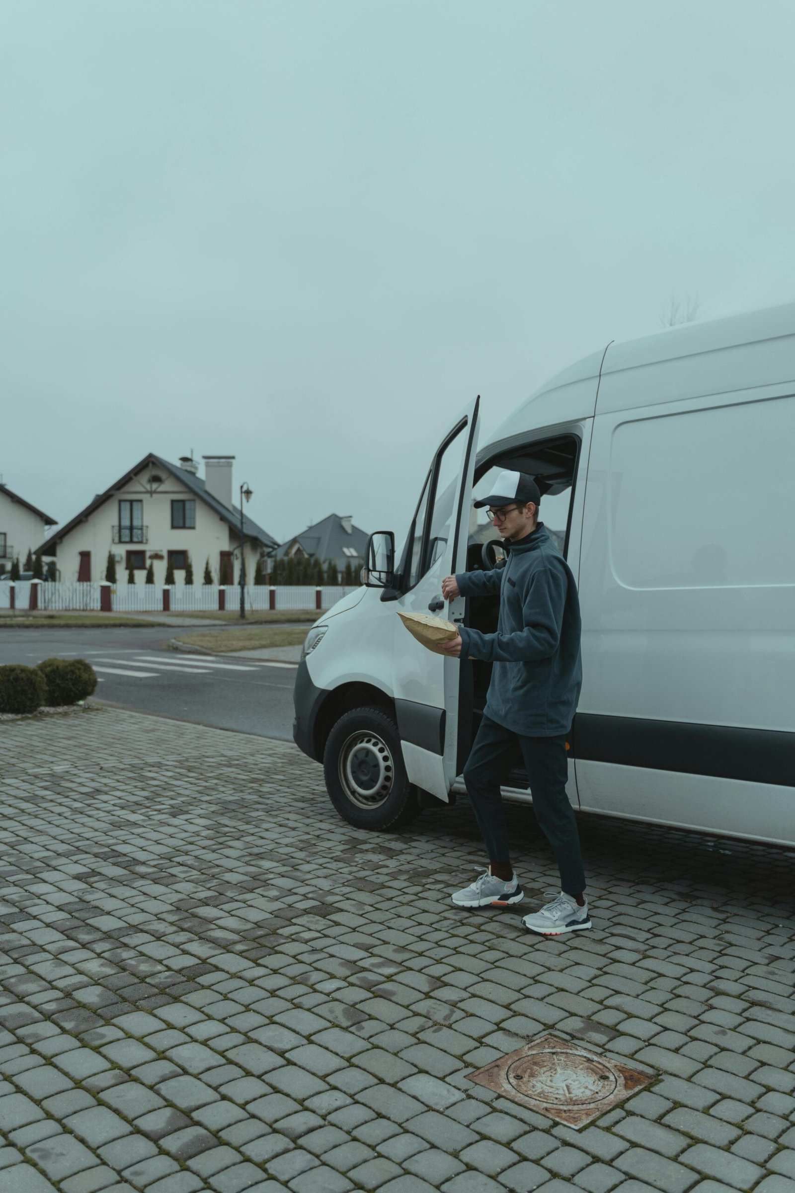 A delivery worker in casual attire carrying a parcel from a white van on a suburban street.